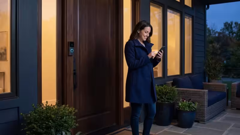 A woman checking her smartphone on a modern front porch with a video doorbell at sunset.