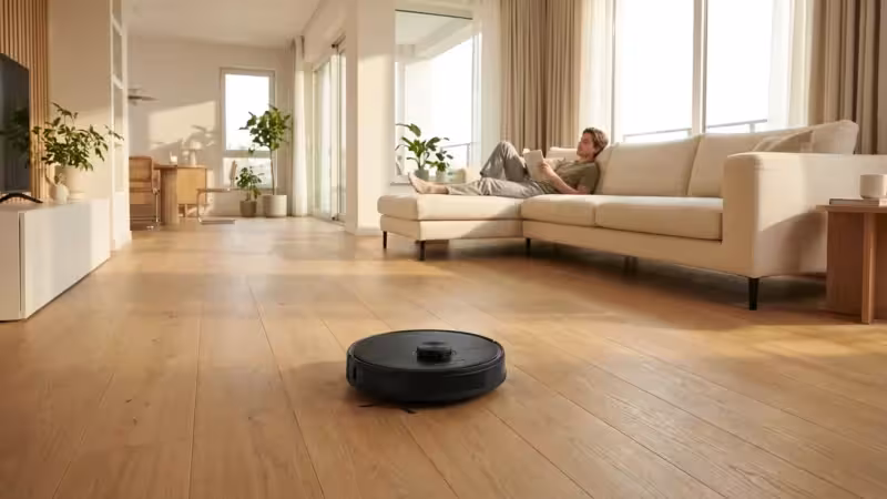 A modern living room with a robot vacuum cleaning a hardwood floor during a sunny afternoon.