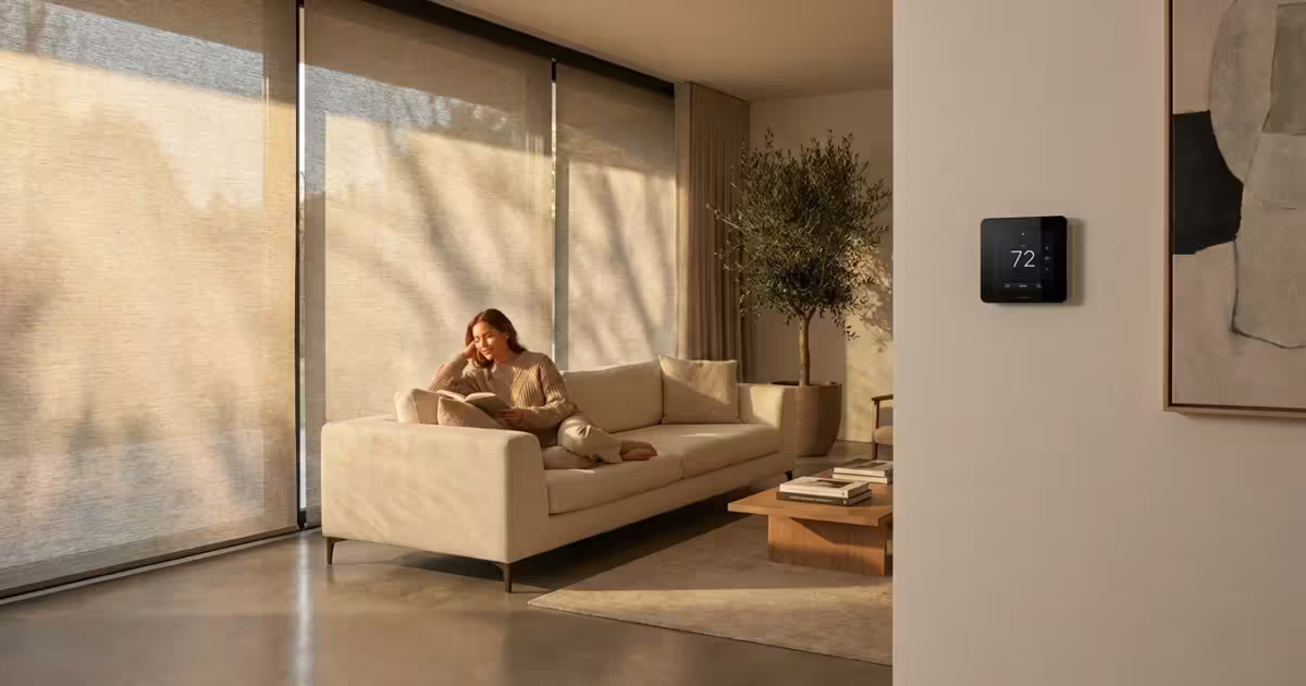 A woman relaxing in a sun-drenched modern living room with automated smart blinds and a wall-mounted thermostat.