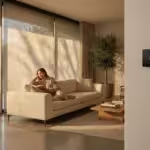 A woman relaxing in a sun-drenched modern living room with automated smart blinds and a wall-mounted thermostat.
