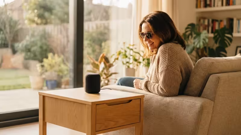 A visually impaired woman smiling while interacting with a smart speaker in a bright, modern living room.
